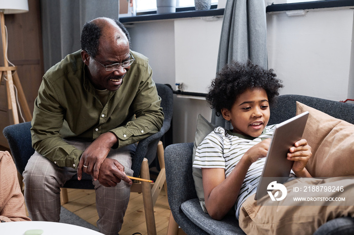 Happy senior African American man discussing online data with his cute grandson pointing at tablet screen while sitting in armchair