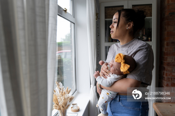 Mother with newborn baby girl looking through window at home