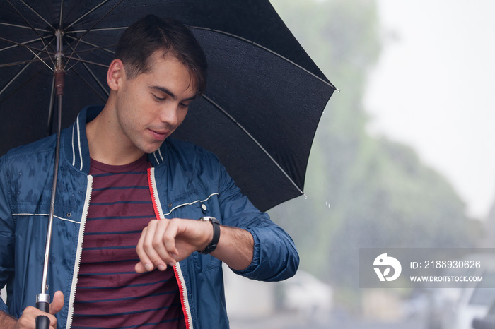 Young man with umbrella checking time on wristwatch in rain