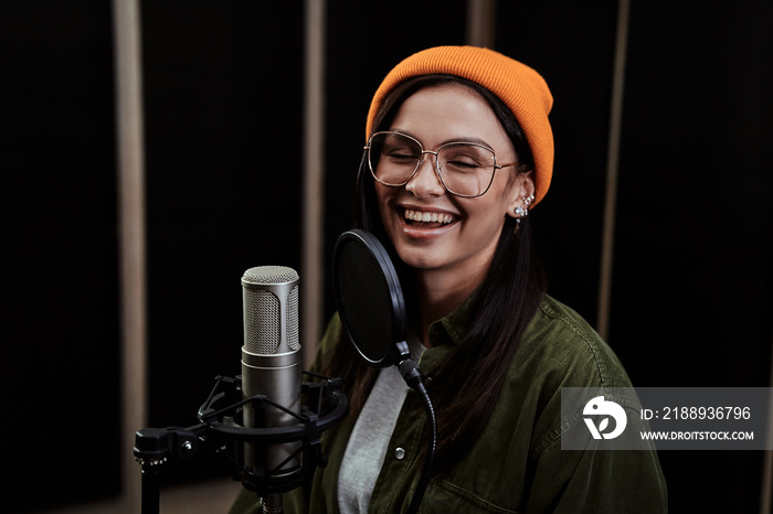 Portrait of cheerful young woman, hip hop artist singing into a condenser microphone while recording