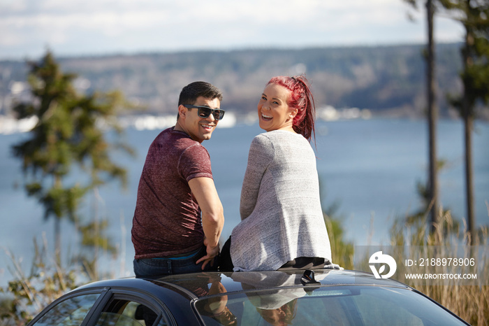 Portrait of young couple sitting on car roof at lakeside looking back