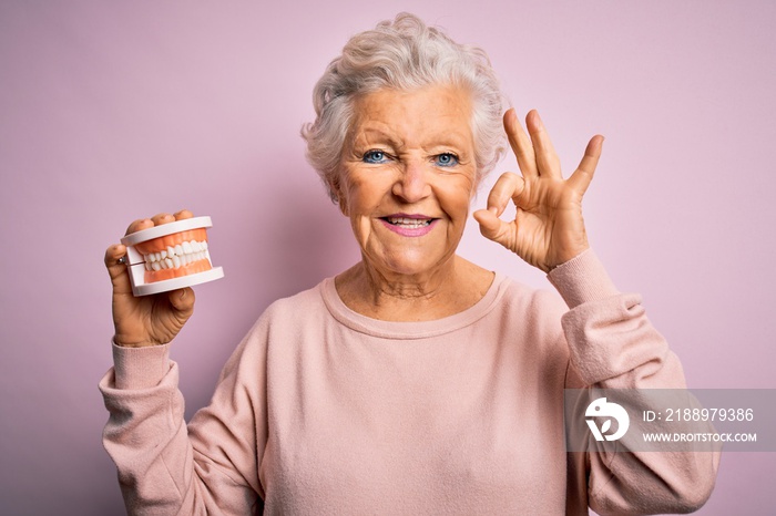 Senior beautiful grey-haired woman holding plastic denture teeth over pink background doing ok sign 