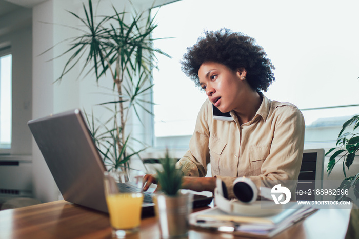 Smiling young African female entrepreneur sitting at a desk in her home office working online with a