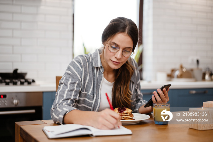 Woman writing down notes and using cellphone while having breakfast