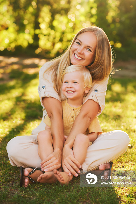 Outdoor sunny portrait of happy young mother hugging adorable toddler boy, family time