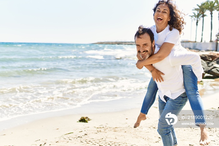 Middle age hispanic man smiling happy holding woman on his back at the beach.