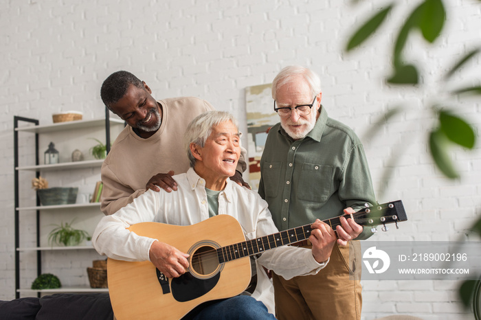 Smiling african american man hugging asian friend playing acoustic guitar at home.