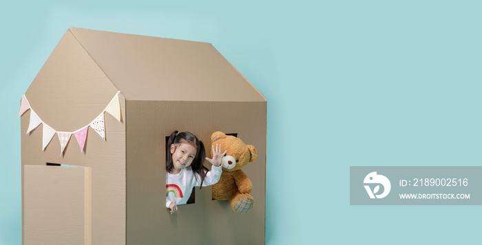 亚洲小女孩带着被隔离在蓝色长横幅上的泰迪熊玩纸板屋