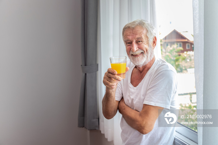 Senior elderly man standing at window in bedroom after waking up in morning with juice, looking came