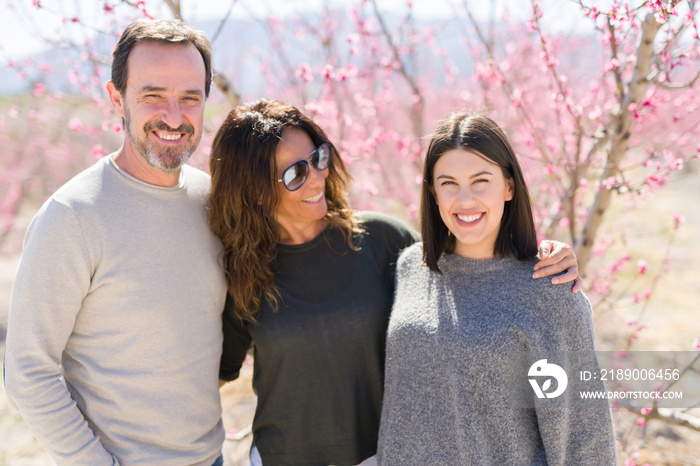 Beautiful family of three smiling cheerful and hugging on peach garden with pink petals enjoying sun