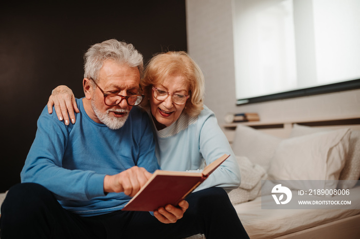 Senior couple is sitting on bed in bedroom and reading from book with red covers. They both are wear