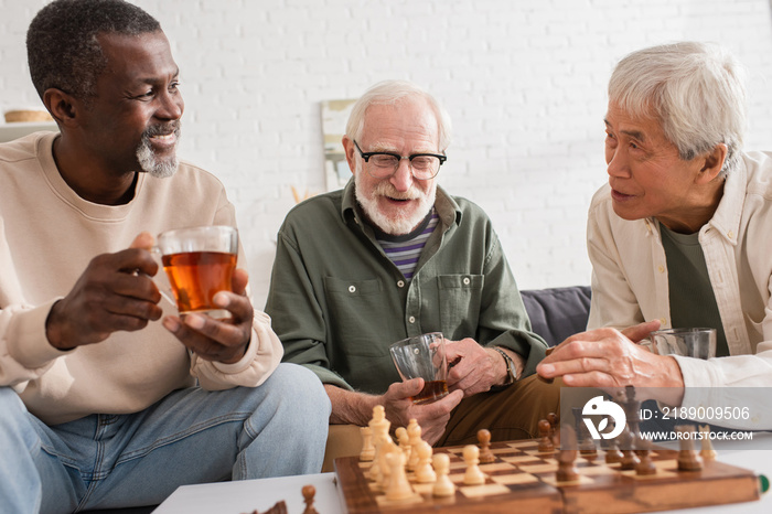 Multicultural friends holding cups of tea near chessboard at home.