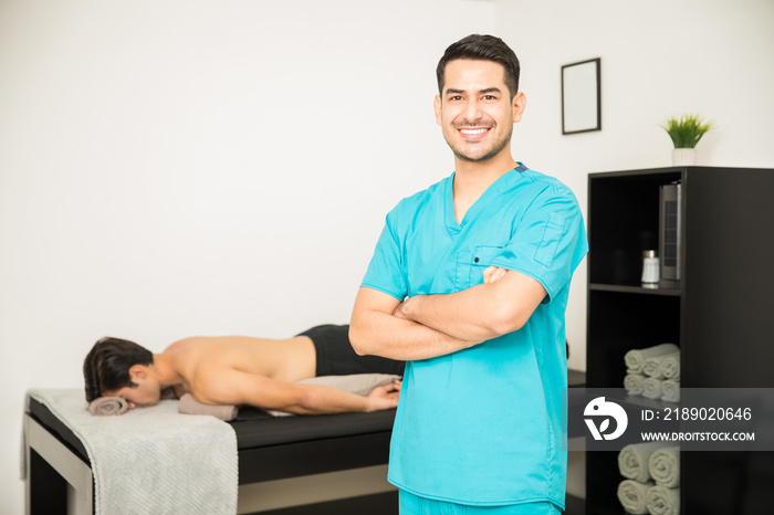 Smiling Physiotherapist Standing Arms Crossed With Patient In Background