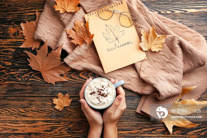 Female hands with hot chocolate in cup, warm sweater and dry leaves on wooden background