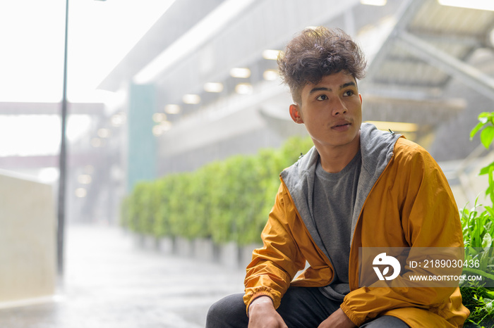 Young Asian man with curly hair thinking and sitting in the city outdoors with rain