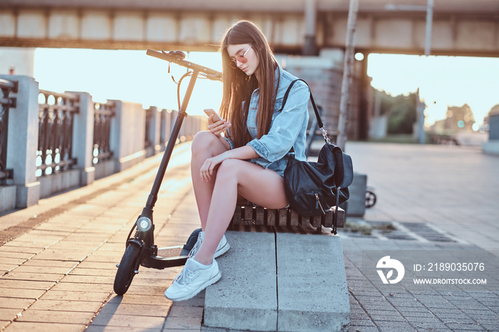 Young attractive woman is chatting by mobile phone while sitting on the bench with her scooter.