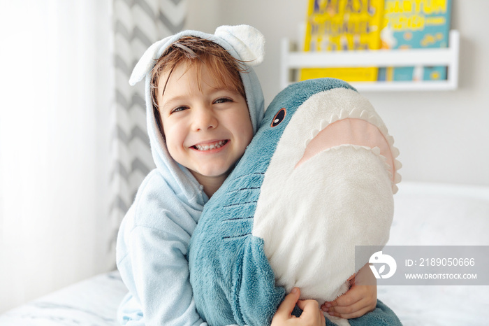 Happy laughing kid boy in soft blue bathrobe after bath play with shark toy on white bed in sunny be