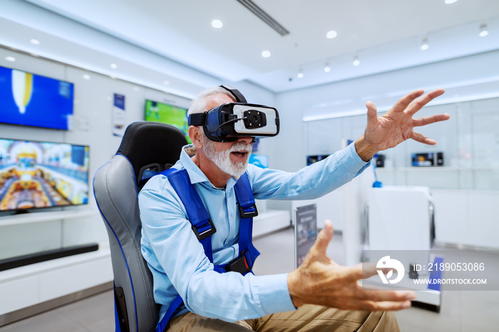 Excited bearded senior man sitting in chair and trying out virtual reality goggles. Tech store inter