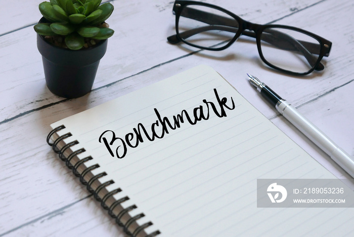 Glasses,plant,pen and notebook written with Benchmark on white wooden background.