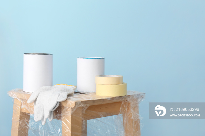 Cans of paint with gloves and tape on stool against color background