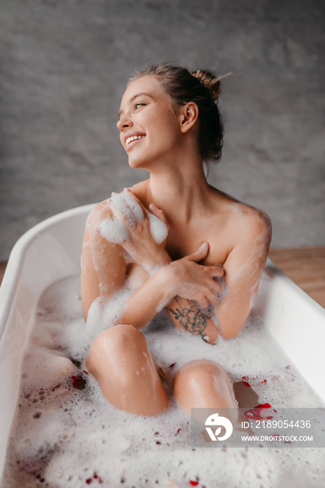 Young sensual woman sitting in hot bubble bath at home, looking aside and smiling, crop, vertical