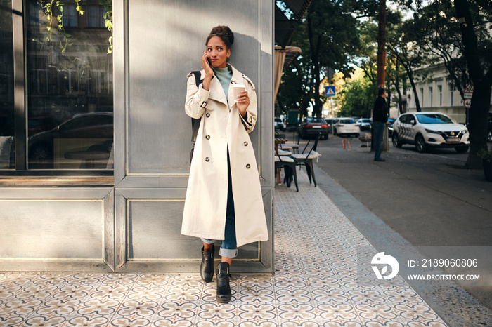 Attractive African American girl in stylish trench coat with coffee to go thinking while talking on 