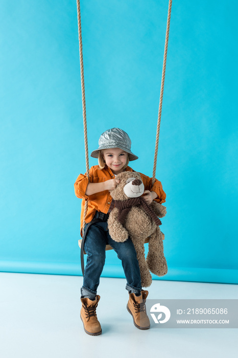 可爱的孩子坐在秋千上，手里拿着蓝色背景的泰迪熊