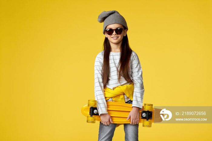 funny little child girl in hat and sunglasses posing with yellow skateboard on yellow studio backgro