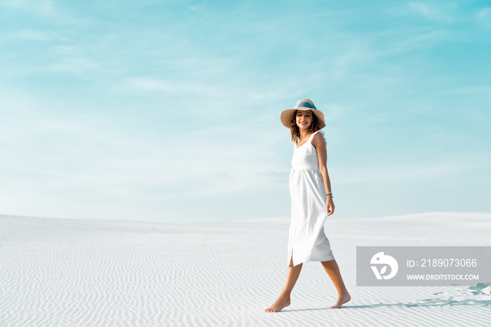 side view of smiling beautiful girl in white dress and straw hat walking on sandy beach with blue sk