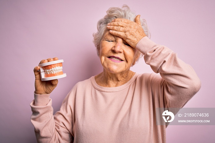 Senior beautiful grey-haired woman holding plastic denture teeth over pink background stressed with 