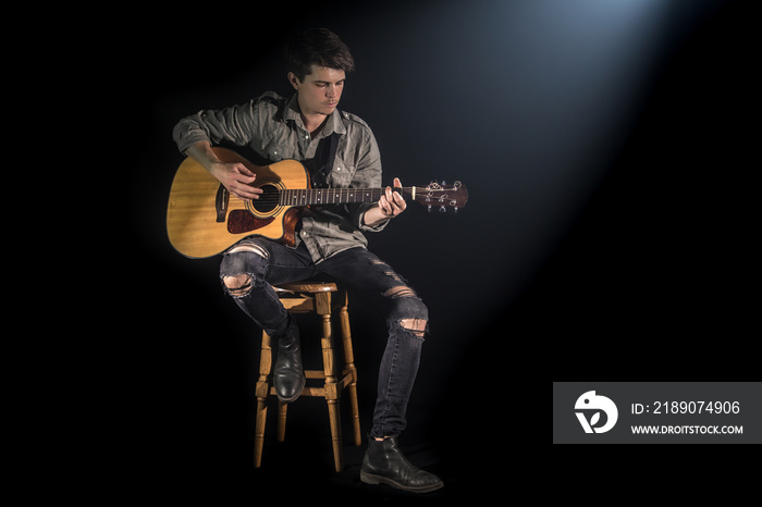 musician playing acoustic guitar, sitting on high chair, black background with beautiful soft light