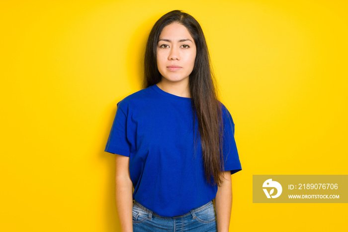 Beautiful brunette woman wearing blue t-shirt over yellow isolated background with serious expressio