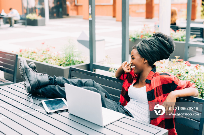 Young beautiful afro black woman sitting outdoor with legs on the table, relaxed, using computer and