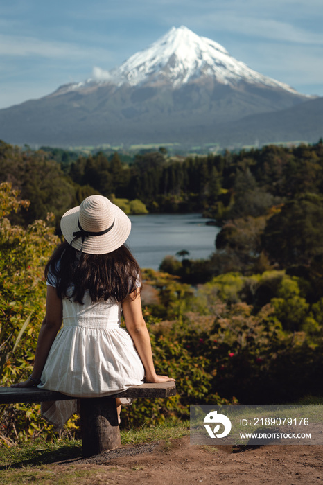 Woman wearing dress sitting on a bench with the beautiful view of Mount Taranaki behind, New Zealand