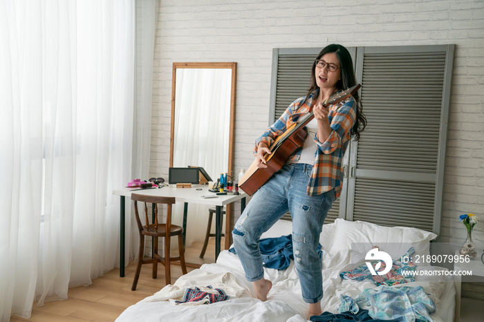 emotional asian woman dressed in shirt holding guitar singing songs jumping on bed. young beautiful 