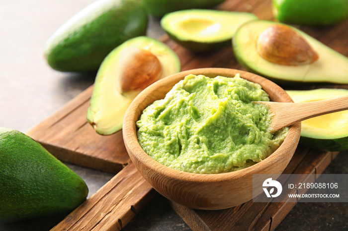 Wooden board with bowl of delicious guacamole and ripe avocados on table