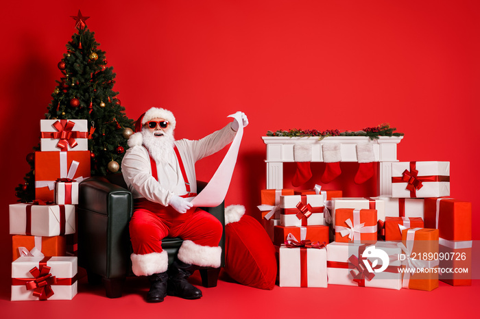 Portrait of his he attractive funky fat cheerful cheery Santa sitting in armchair reading long paper