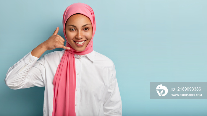 Happy young Muslim female model gestures with fingers, says call me, smiles broadly, wears pink hija