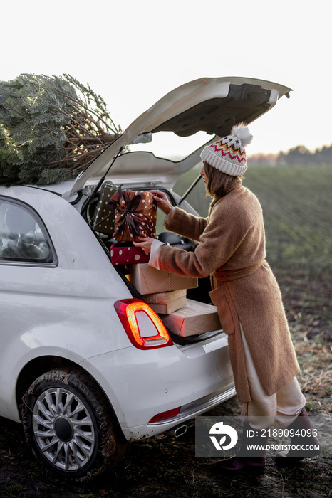 Woman packing gifts into the car with Christmas tree on a rooftop on nature at dusk. Getting ready f