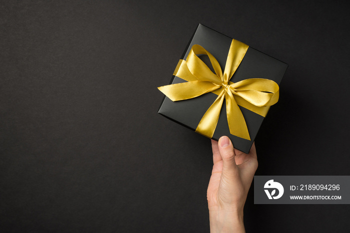 First person top view photo of hand giving black giftbox with golden satin ribbon bow on isolated bl