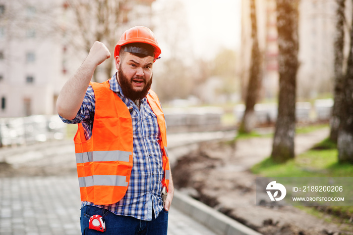 Portrait of brutal angry beard worker man suit construction worker in safety orange helmet against p