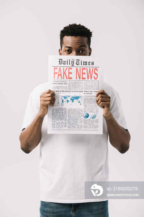 curly african american man covering face while holding newspaper with fake news lettering isolated o