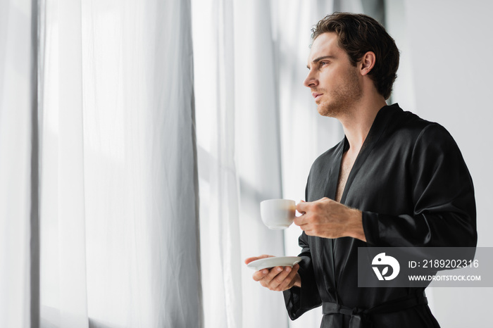 Young man in black robe holding coffee cup and saucer near curtains at home