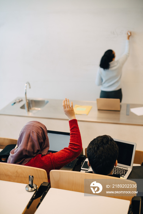 High angle view of teenage girl raising hand while studying with classmate in university lecture