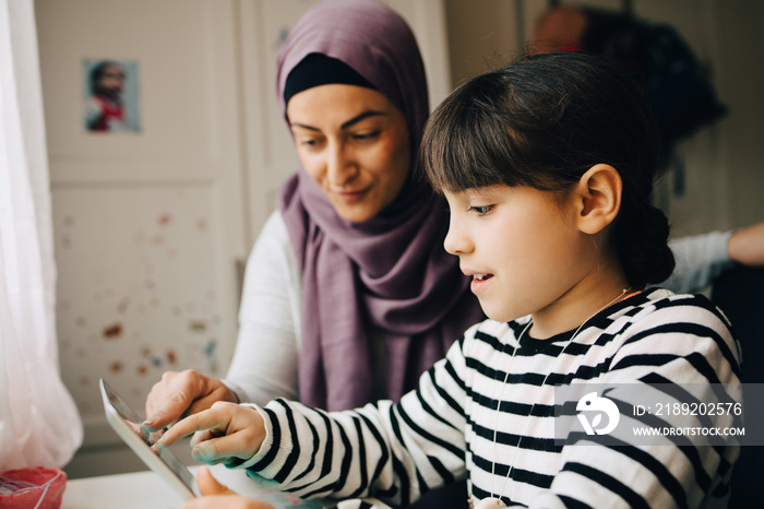 Girl sitting with mother learning homework from digital tablet at home