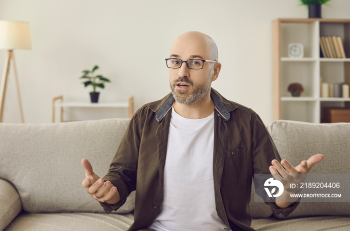 Bald bearded adult man in glasses sitting on sofa, looking at camera, talking and gesturing. Mature 