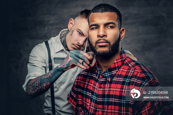 Tattooed barber cuts the beard to a Black bearded male.