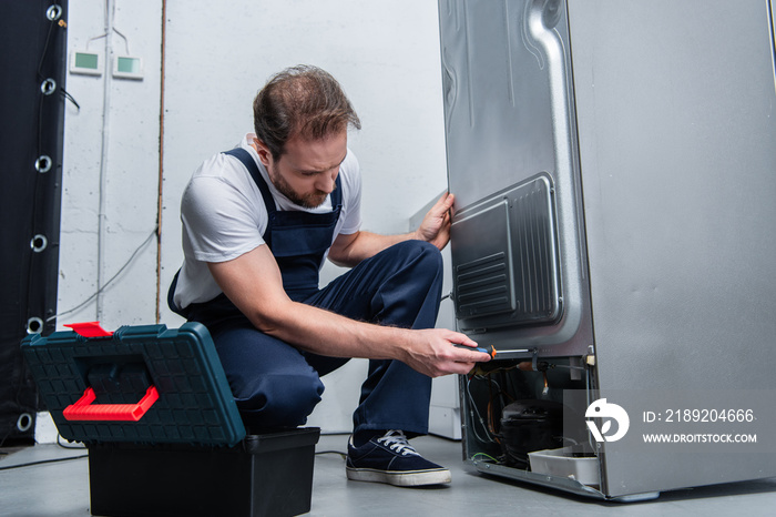 adult bearded repairman in working overall fixing refrigerator by screwdriver in kitchen