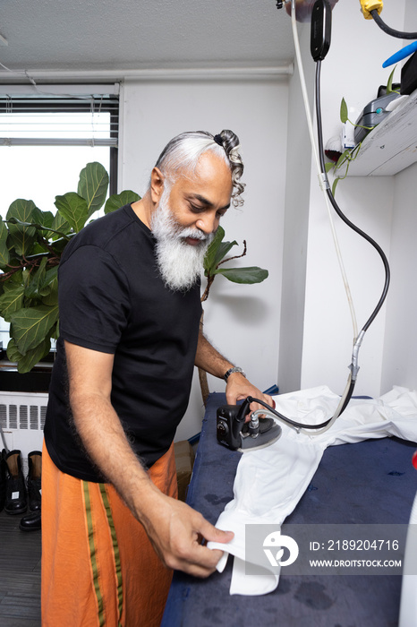 gray hair bearded man wearing skirt and ironing a white shirt