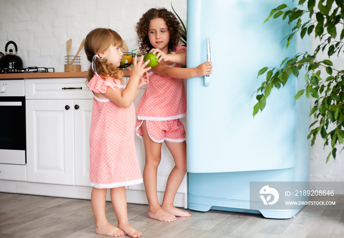 两个小女孩在厨房里吃水果。孩子们在冰箱旁吃有益健康的食物。